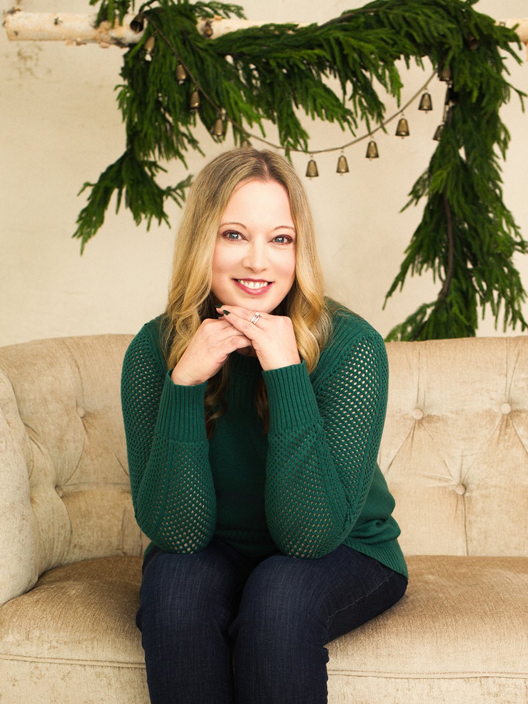 Amanda Weinraub in a green sweater, smiling on a velvet settee beneath a garland of greenery and brass bells