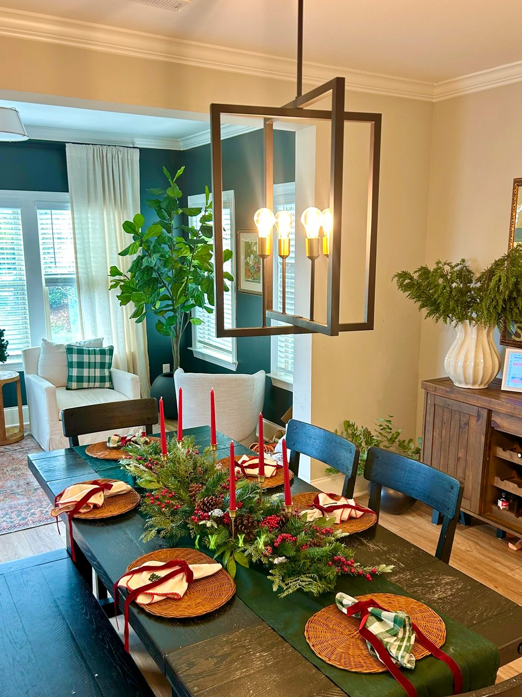 A dining room table set for the holidays with a greenery runner, red candles, and woven chargers