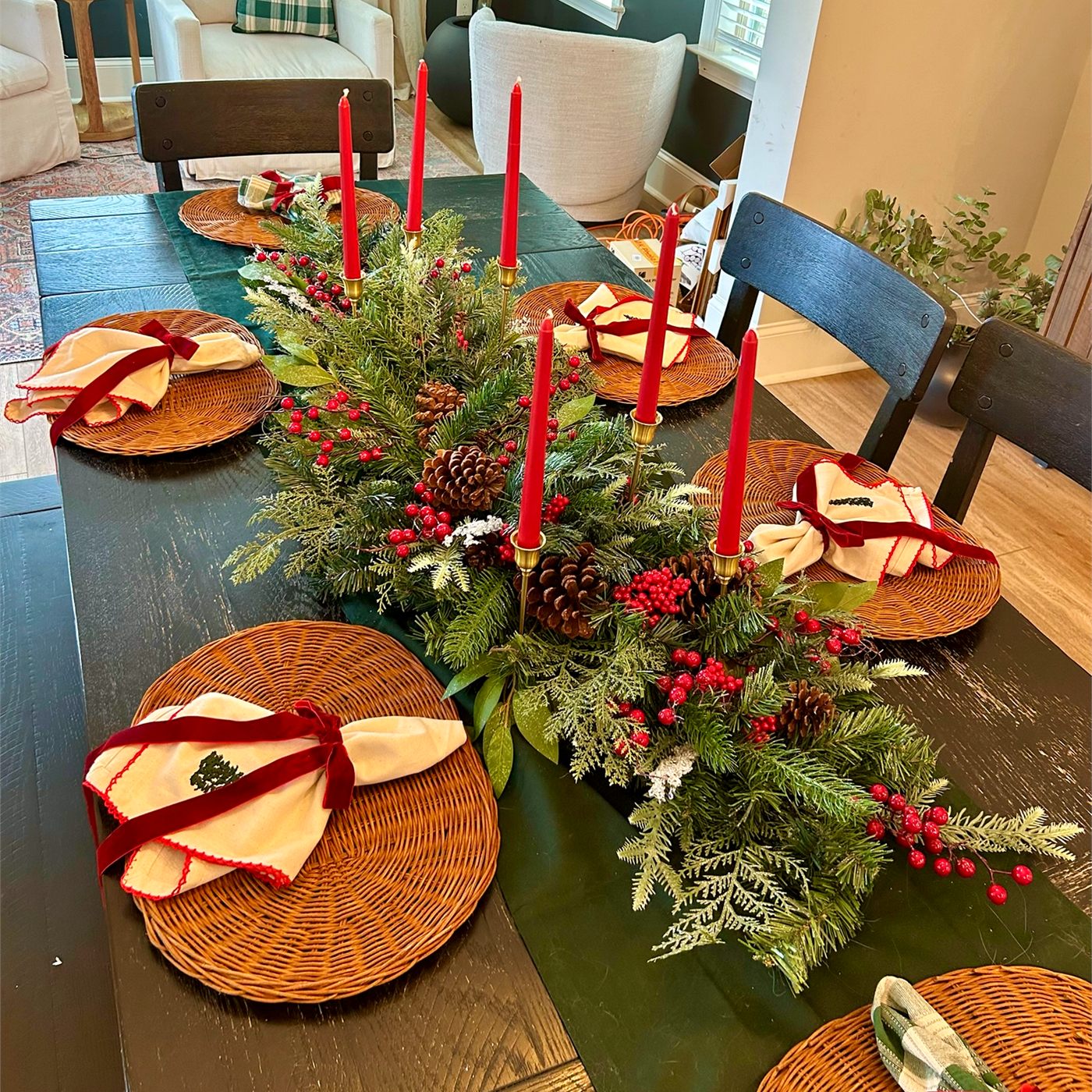 A holiday dining table with a greenery centerpiece, red taper candles, and woven chargers
